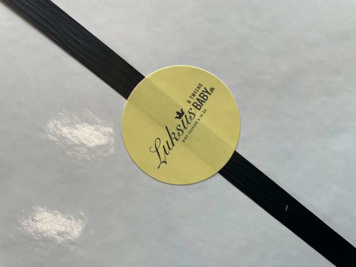 Et tyndt sort bånd vikler sig om en hvid æske, fastgjort med et rundt gult klistermærke. Klistermærket har "Luksusbaby Danmark" skrevet i kursiv sammen med ekstra tekst og et lille kronedesign. Lysrefleksioner er synlige på overfladen af Gaveindpakningen.