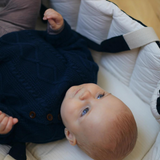 En baby med lys hud og blå øjne ligger på et hvidt tæppe og kigger op. Babyen er iført en marineblå striktrøje med knapper foran. Babyen er omgivet af quiltet polstring i en Copenhagen Colors Babylift og Legetæppe Sort m. Creme Solid Stripe, med en ergonomisk håndtag til nem at bære fra COPENHAGEN COLORS.