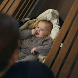 En baby iført en grå sweater ligger i en trækrybbe og kigger op med et nysgerrigt udtryk. Tremmesengen, foret med en blød, beige pude, har et ergonomisk designet håndtag for nem bevægelse. The Copenhagen Colors Babylift og Legetæppe Sort m. Creme Solid Stripe fra COPENHAGEN COLORS ses i baggrunden. En voksen er delvist synlig i forgrunden og stirrer på babyen.
