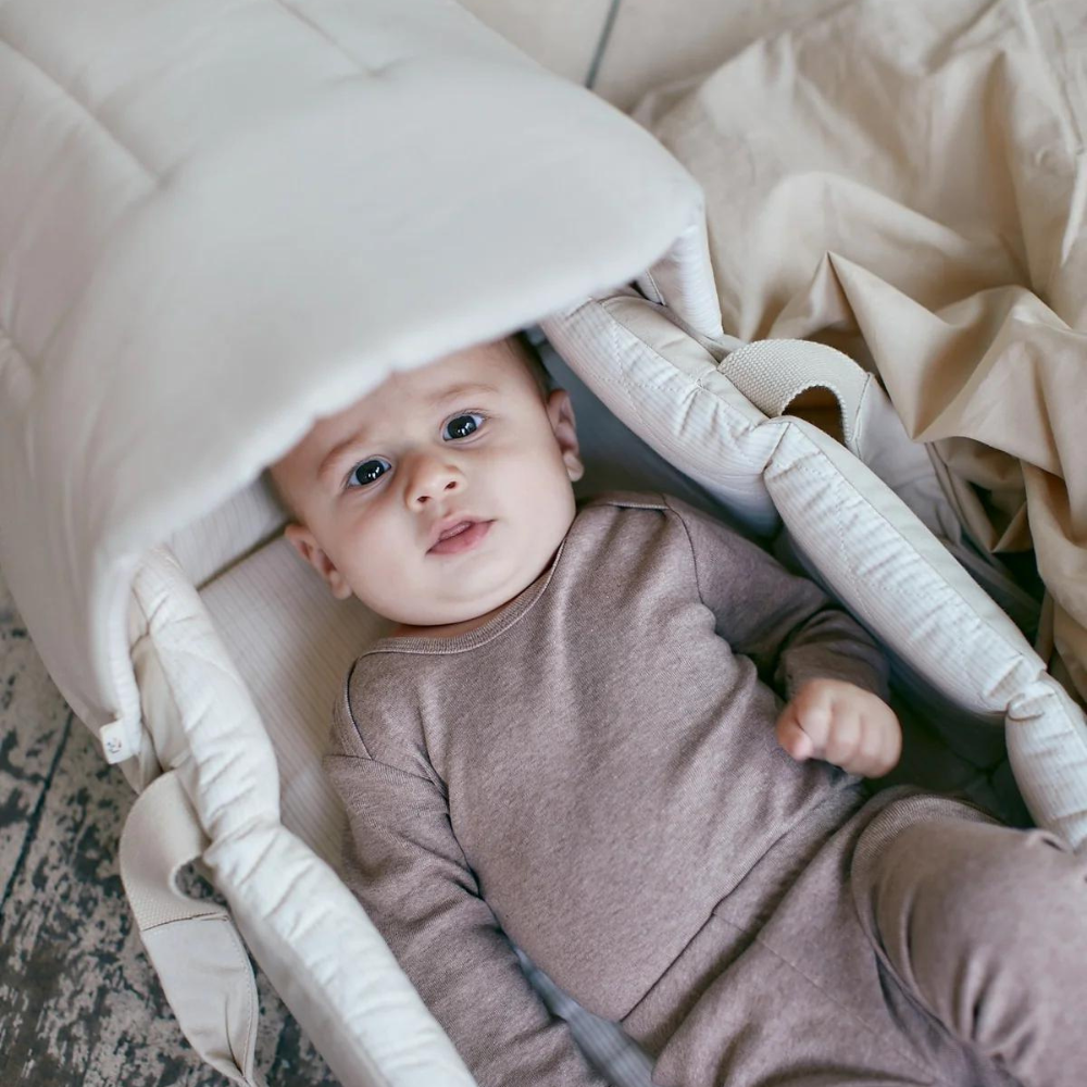 En baby ligger i en blød, polstret Copenhagen Multifunktionel Kaleche til Babylift Creme Solid m. Creme Stripe by COPENHAGEN COLORS på gulvet lavet af økologisk bomuld. Babyen er iført et beige outfit og har et vågent udtryk og kigger op i kameraet. Bæreren er polstret med genanvendt polyester og delvist dækket med et beige tæppe. Baggrunden viser et tekstureret gulv og mere stof.