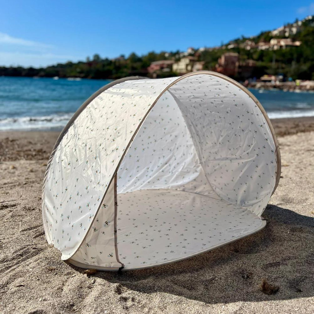 Et Vanilla COPENHAGEN Pop-Up Telt UV50+ Honeybee Print med et subtilt honningbimønster er sat op på en sandstrand. Kystlinjen og de blide bølger er synlige i baggrunden sammen med en klar blå himmel og fjerne huse beliggende blandt grønne områder. Teltet, med UV-beskyttelse og en opbevaringstaske, er delvist åbent og ubeboet.